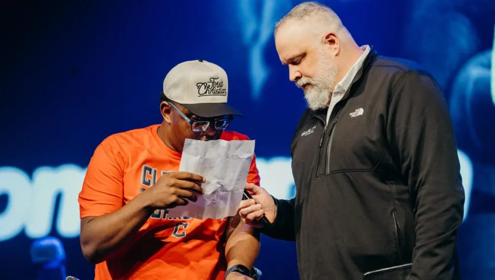 A young man reads aloud on stage while a pastor stands beside him